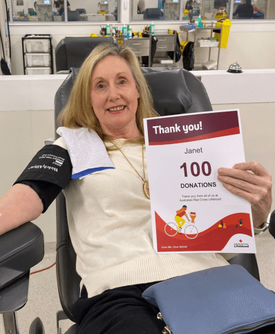 Janet has blonde hair and is wearing a yellow t-shirt. She sits in a chair at Lifeblood holding a certificate in her left hand and her right arm is stretched out with a tourniquet.